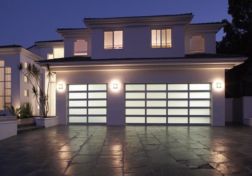A large modern home with two aluminum garage doors with blurred privacy windows. Dark outside, lights on inside.