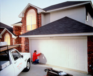 Overhead Door Company garage door technician inspecting large white garage door on brick home.